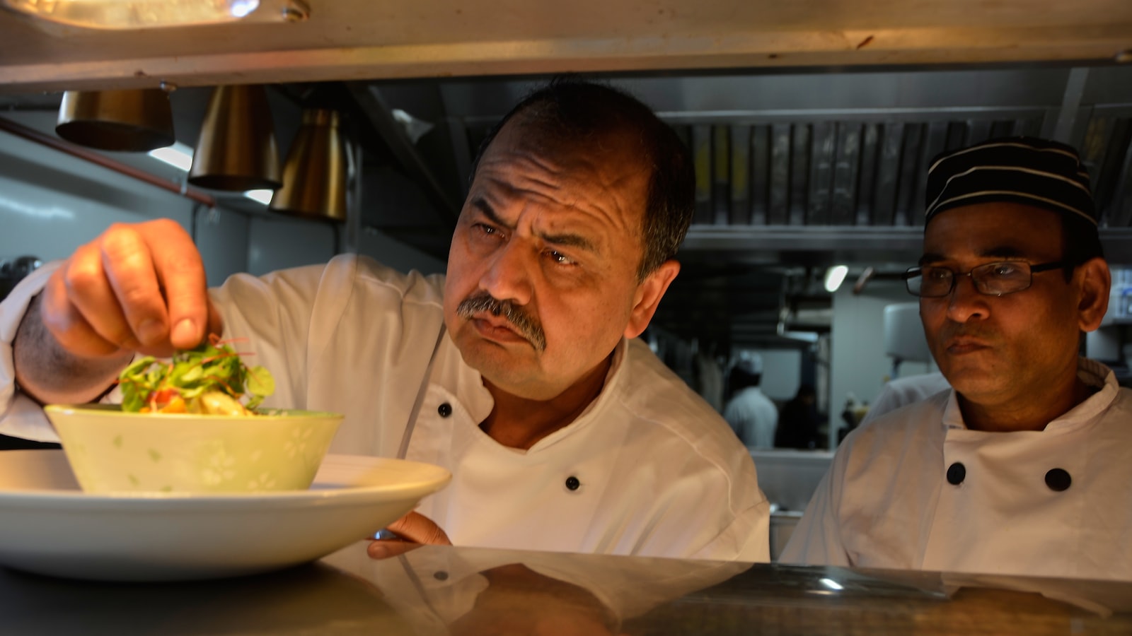A head chef finishing a plate at the pass while a cook watches, restaurant leadership shown by setting the standard